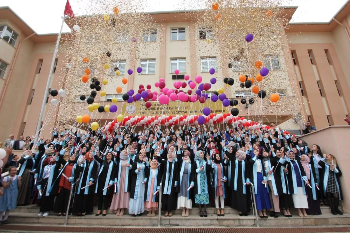 Okul binası önünde turkuaz bantlı kiralık kep-cübbelerle ortaokul 8. sınıf mezuniyet töreni