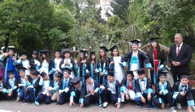 Okul bahçesinde turkuaz bantlı kiralık kep-cübbelerle ilkokul mezuniyet töreni toplu fotoğrafı