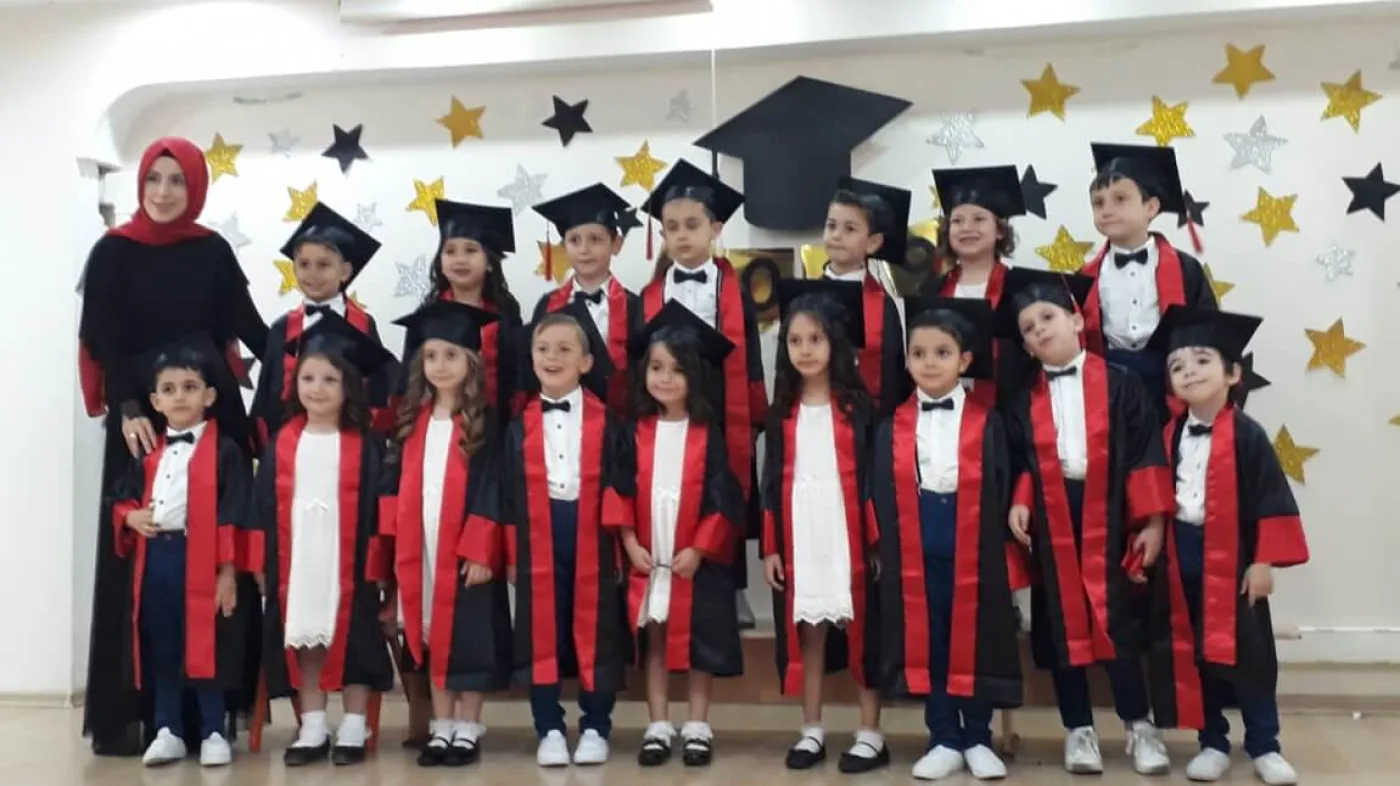 Sahnede kırmızı bantlı kiralık kep-cübbelerle ilkokul mezuniyet töreni